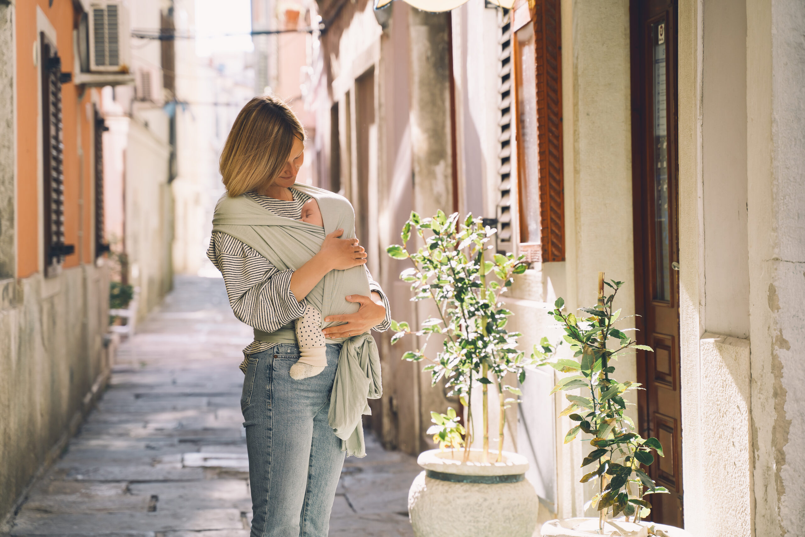 Babywearing. Mother and baby walking in town. Baby in wrap carrier. Young woman carrying little child in baby sling in mint color. Concept of green parenting, natural motherhood, postpartum period.