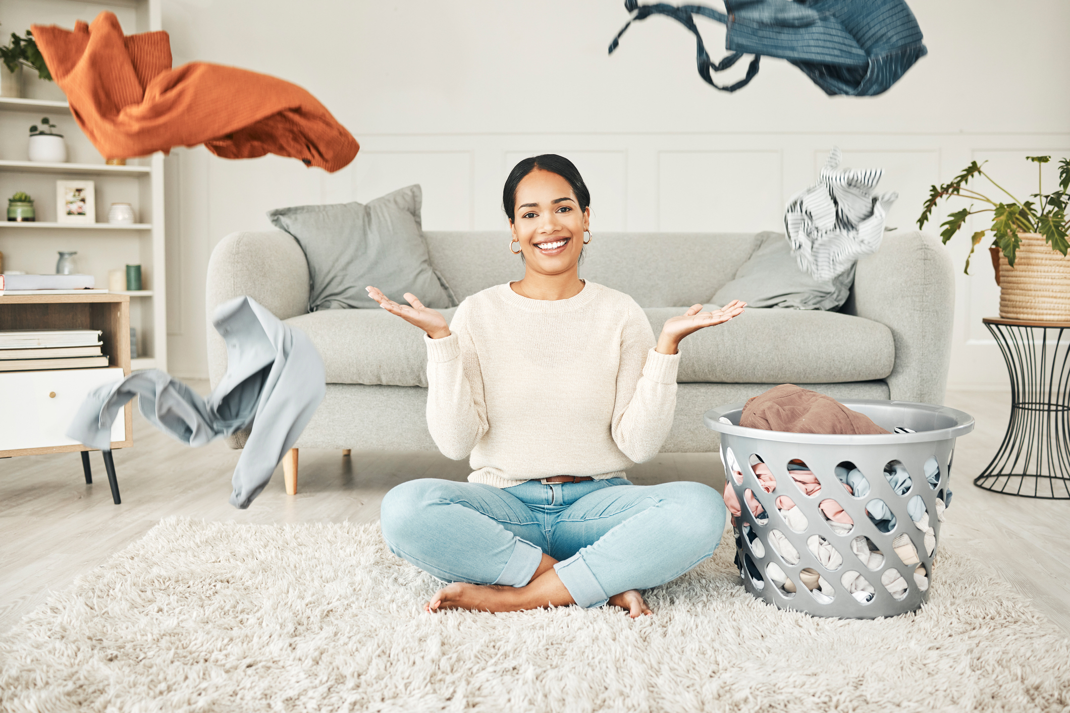 Portrait of a cleaning, carefree female cleaner throwing clothing in the air. Happy, smiling and young woman doing laundry, washing clothes and sitting in a messy living room at home.