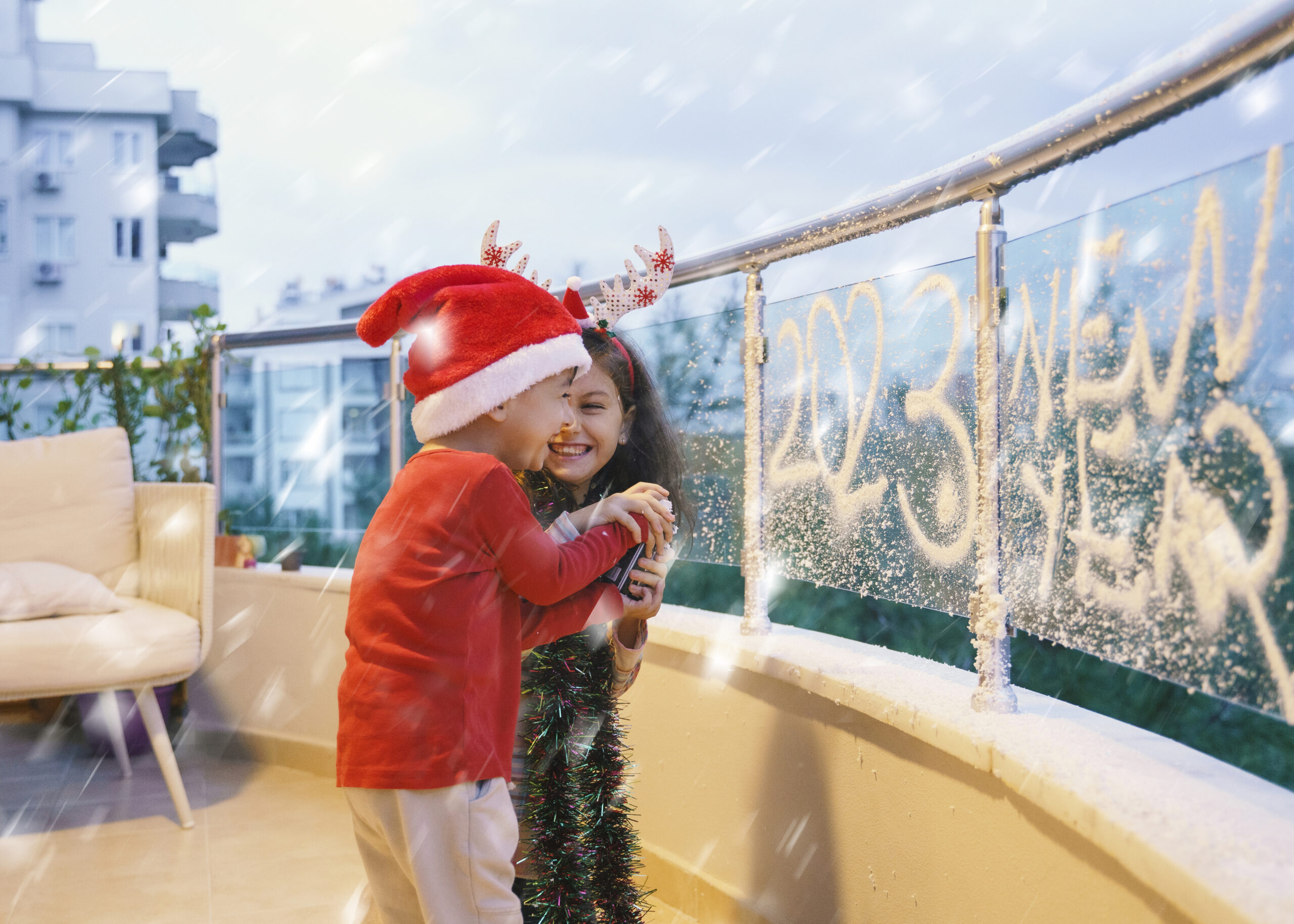 Happy children (brother and sister) spraying fake snow on balcony glass, Christmas decoration