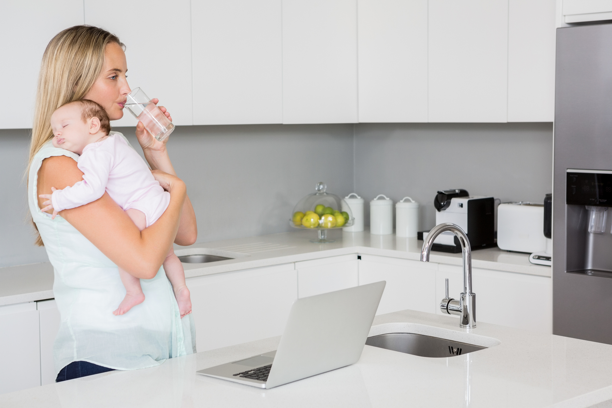 Mother drinking water while carrying baby in kitchen