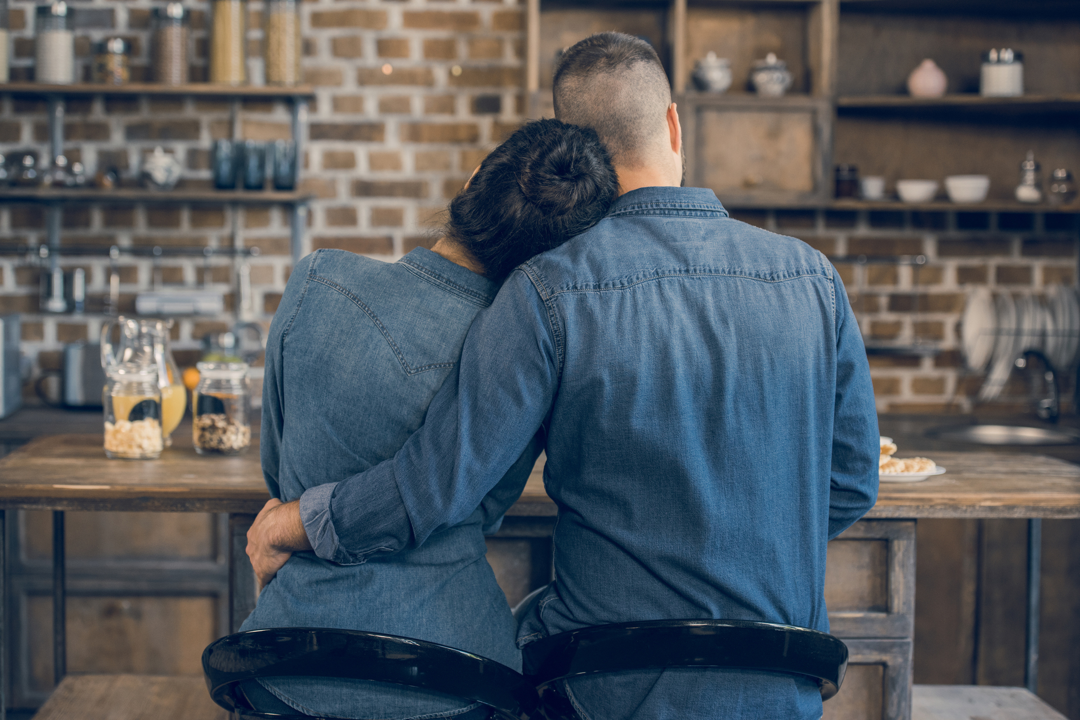 Back view of young couple in denim shirts sitting embracing at wooden table and having breakfast