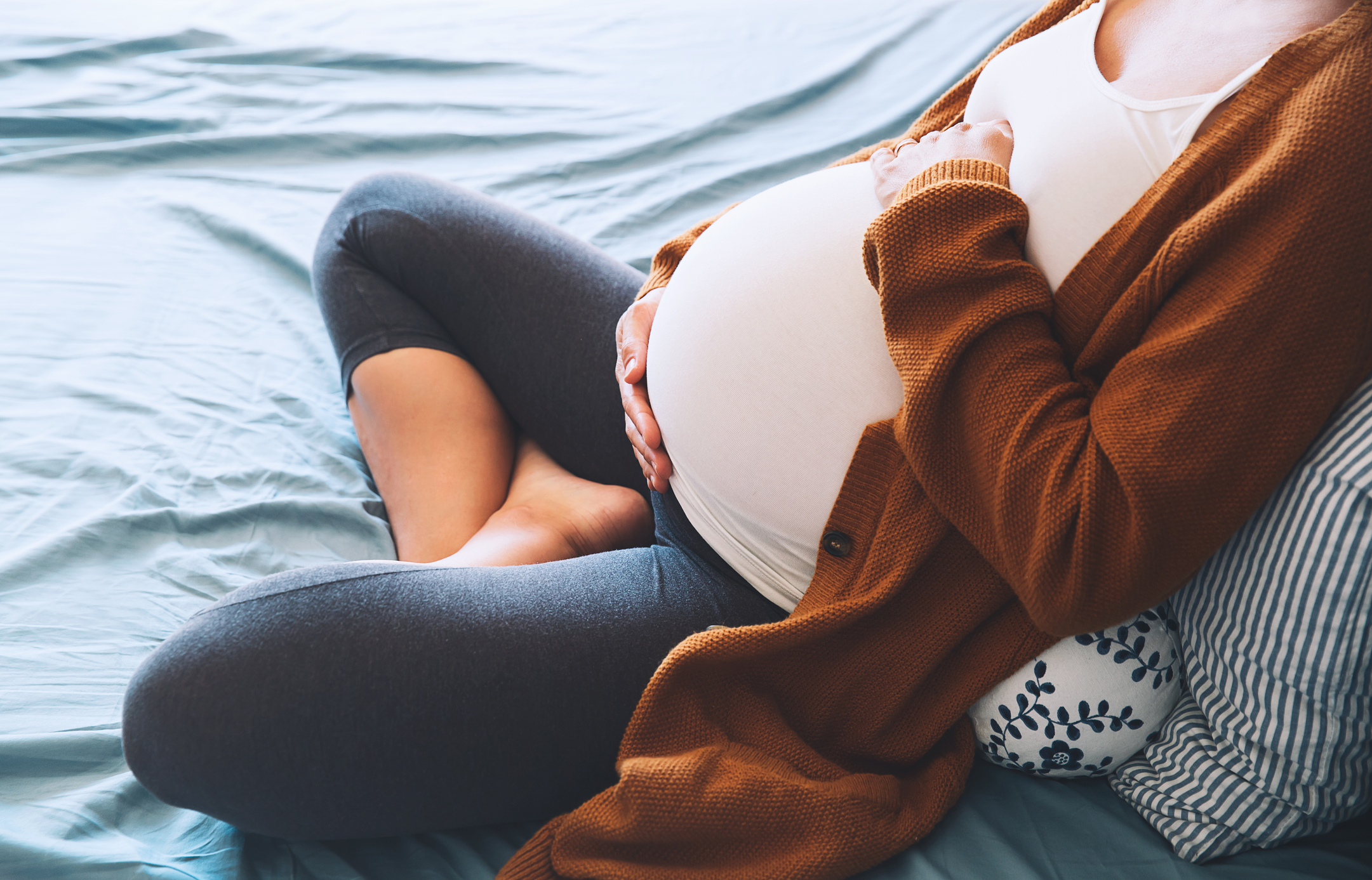 Beautiful pregnant woman sitting at bed and holds hands on belly in bedroom at home.