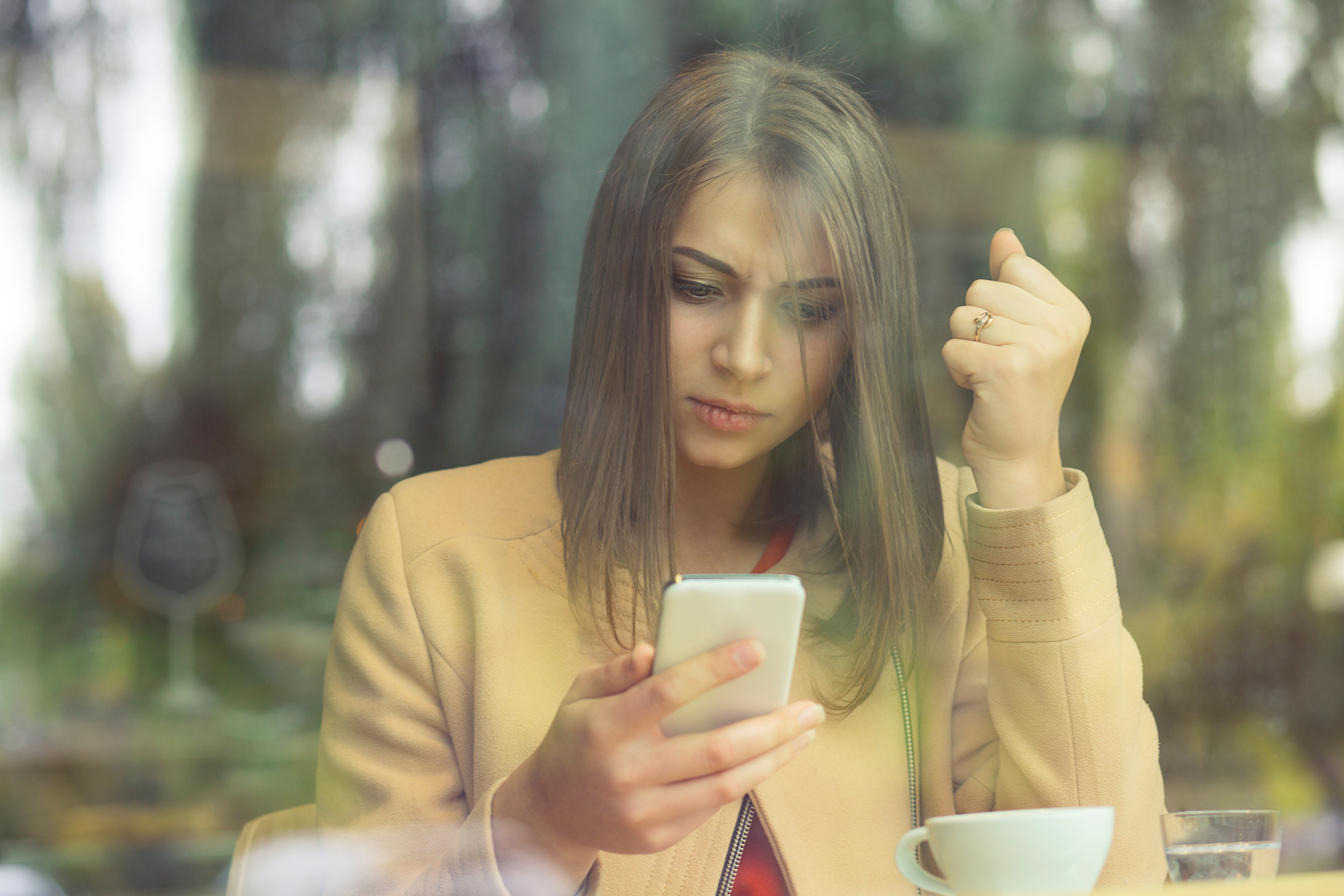 Upset stressed woman holding cellphone angry with message she received fist up on phone isolated coffee shop background. Sad looking human face expression emotion feeling body language