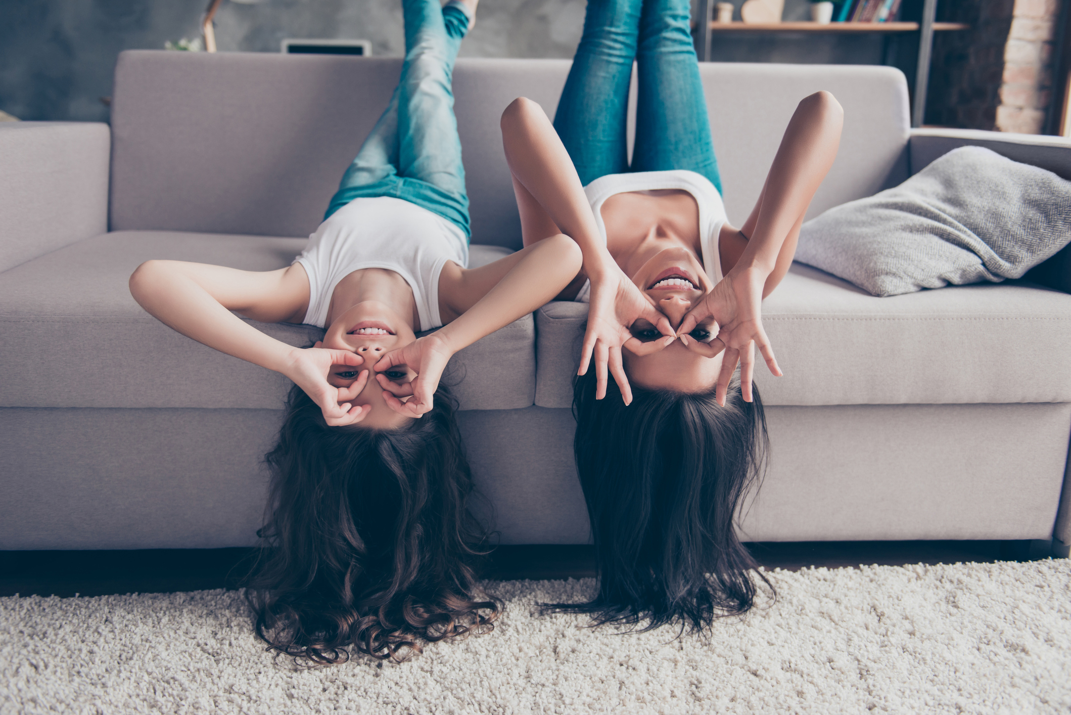 Lovely cheerful happy mother and playful little daughter with long hair are lying upside down on a couch and making binoculars with their fingers