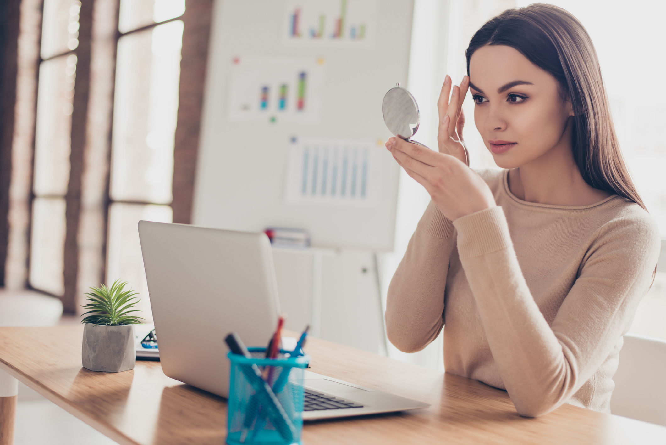 Portrait of attractive charming pretty tender gentle fresh carefree stunning elegant gorgeous employer correcting doing make up looking in mirror touching brows sitting at modern light office