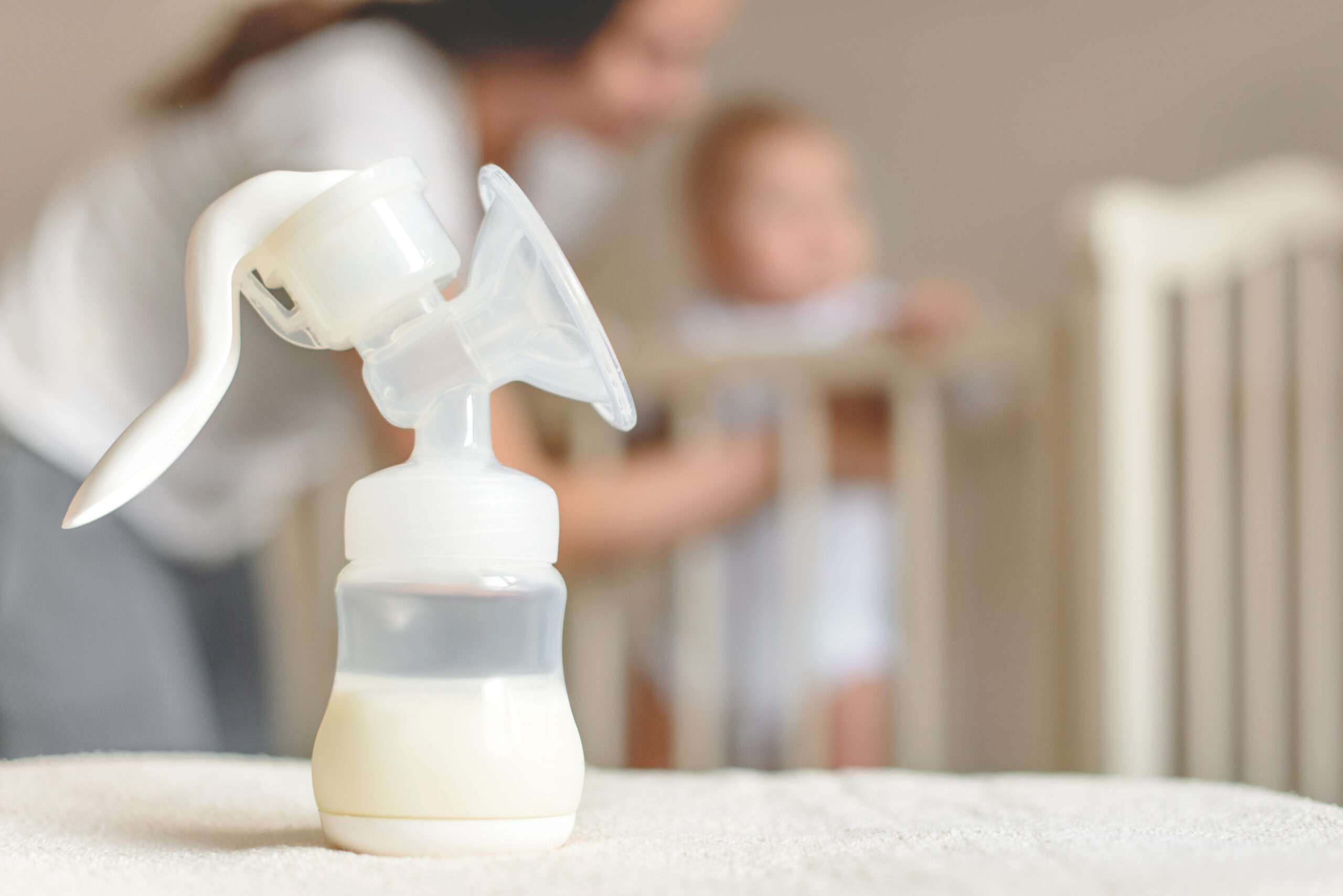 Manual breast pump and bottle with breast milk on the background of mother and baby near the baby's bed.