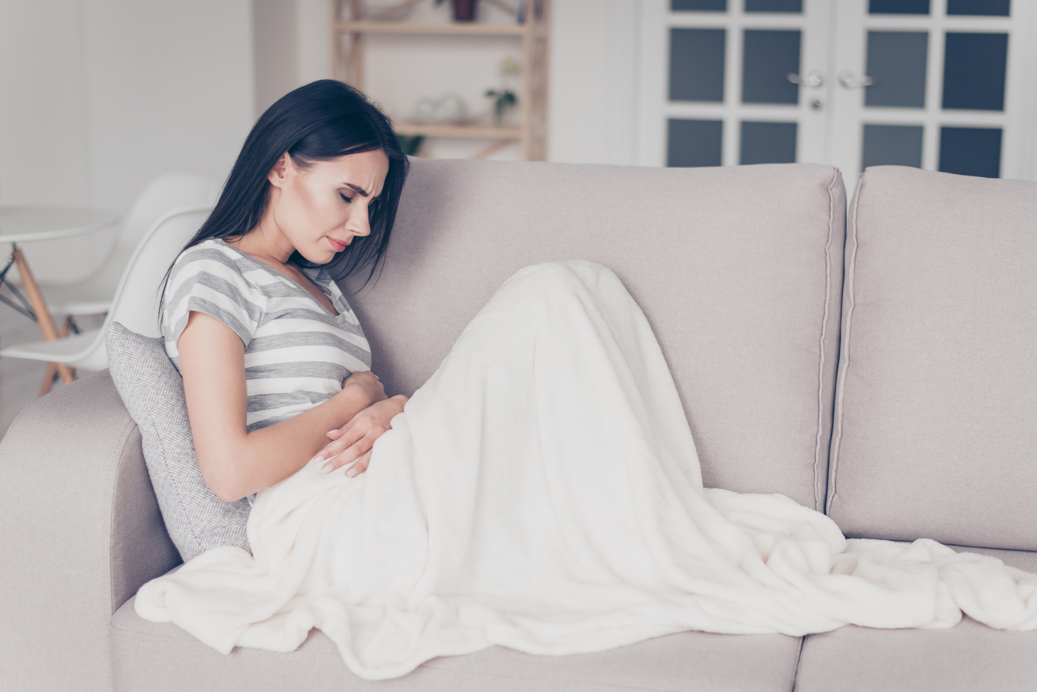 sad beautiful woman sitting couch and feel spasm and symptoms of pms while touch belly with coverlet