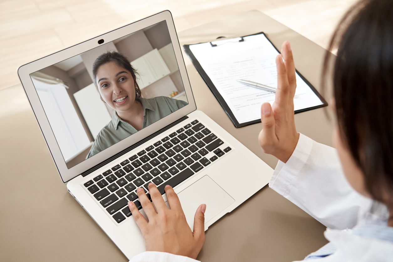 Female doctor gp consulting indian woman patient make online webcam video call on laptop screen. Telemedicine conference remote computer virtual visit in meeting. Over shoulder videocall view.