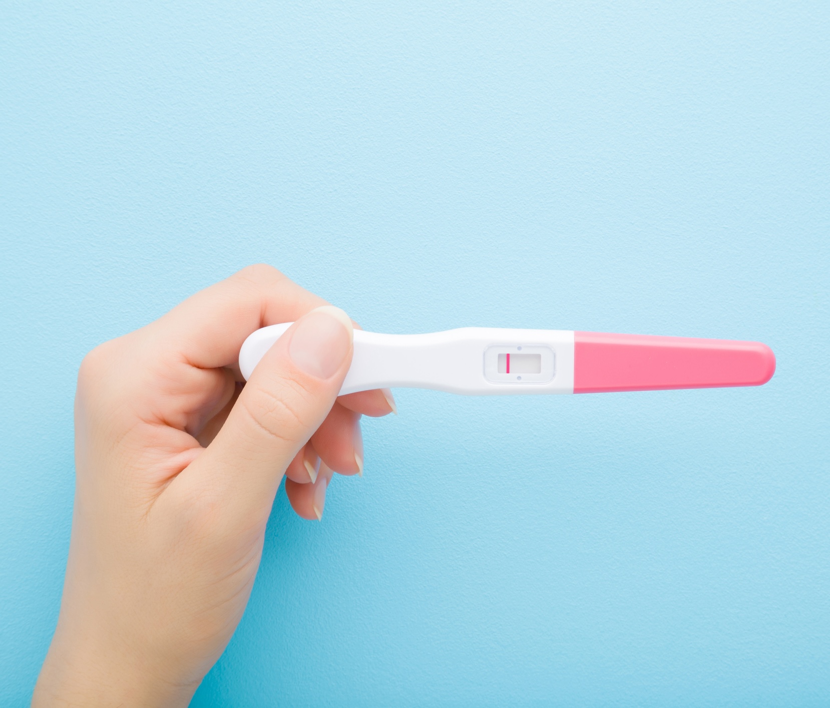 Young adult woman hand holding pregnancy test with one stripe on light blue table background. Pastel color. Negative result. Closeup. Top down view.