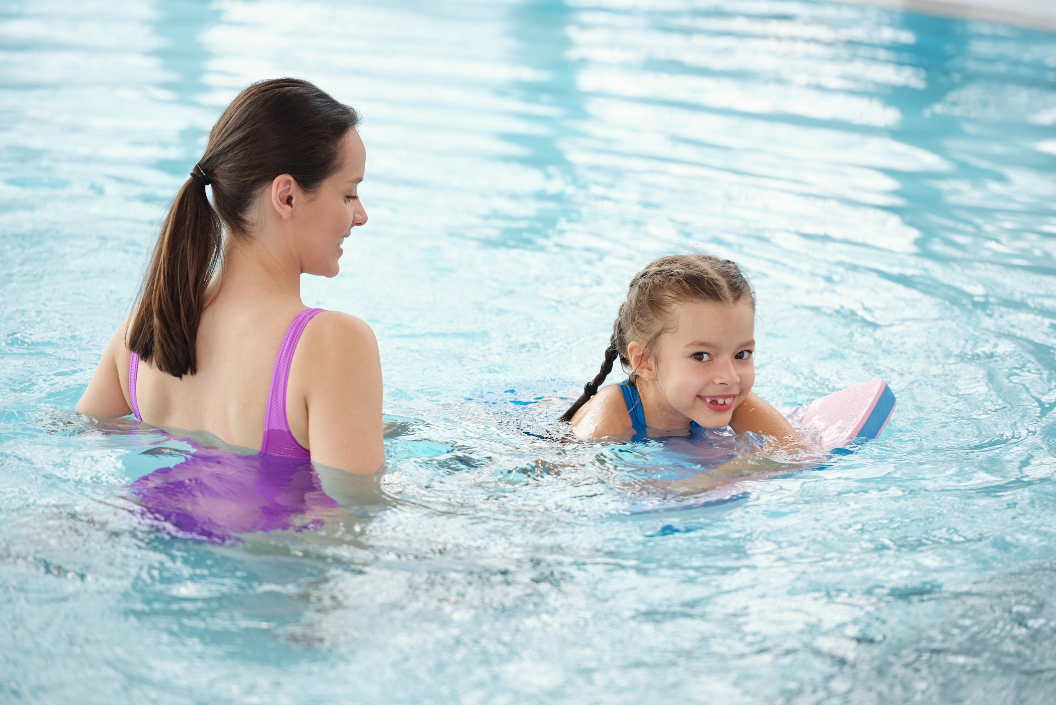 Happy young brunette woman helping adorable little daughter to swim in pool