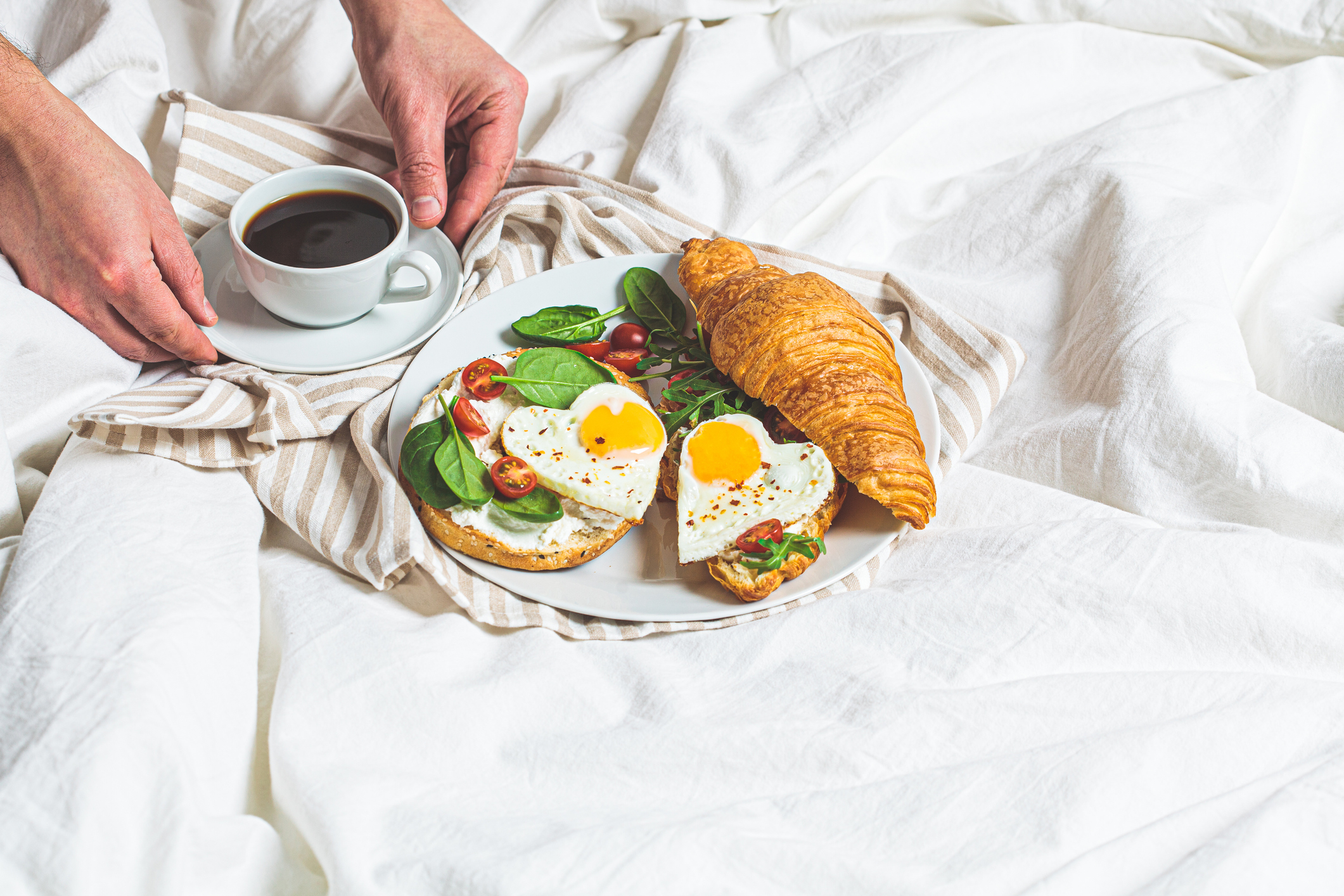 Valentine's day concept. Breakfast in bed on Valentine's Day. Sandwiches with croissant, bagel, cream cheese and fried eggs hearts on white bed background, copy space.