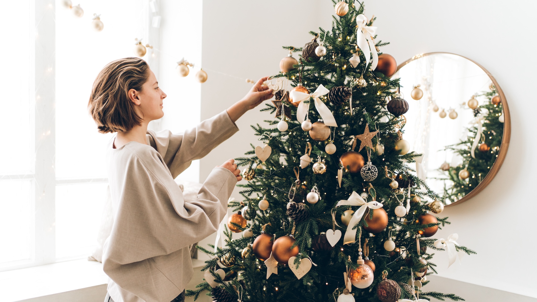 Young woman is adjusting her hair and decorating Christmas tree with many different decorations and festive garland. New Year celebration concept