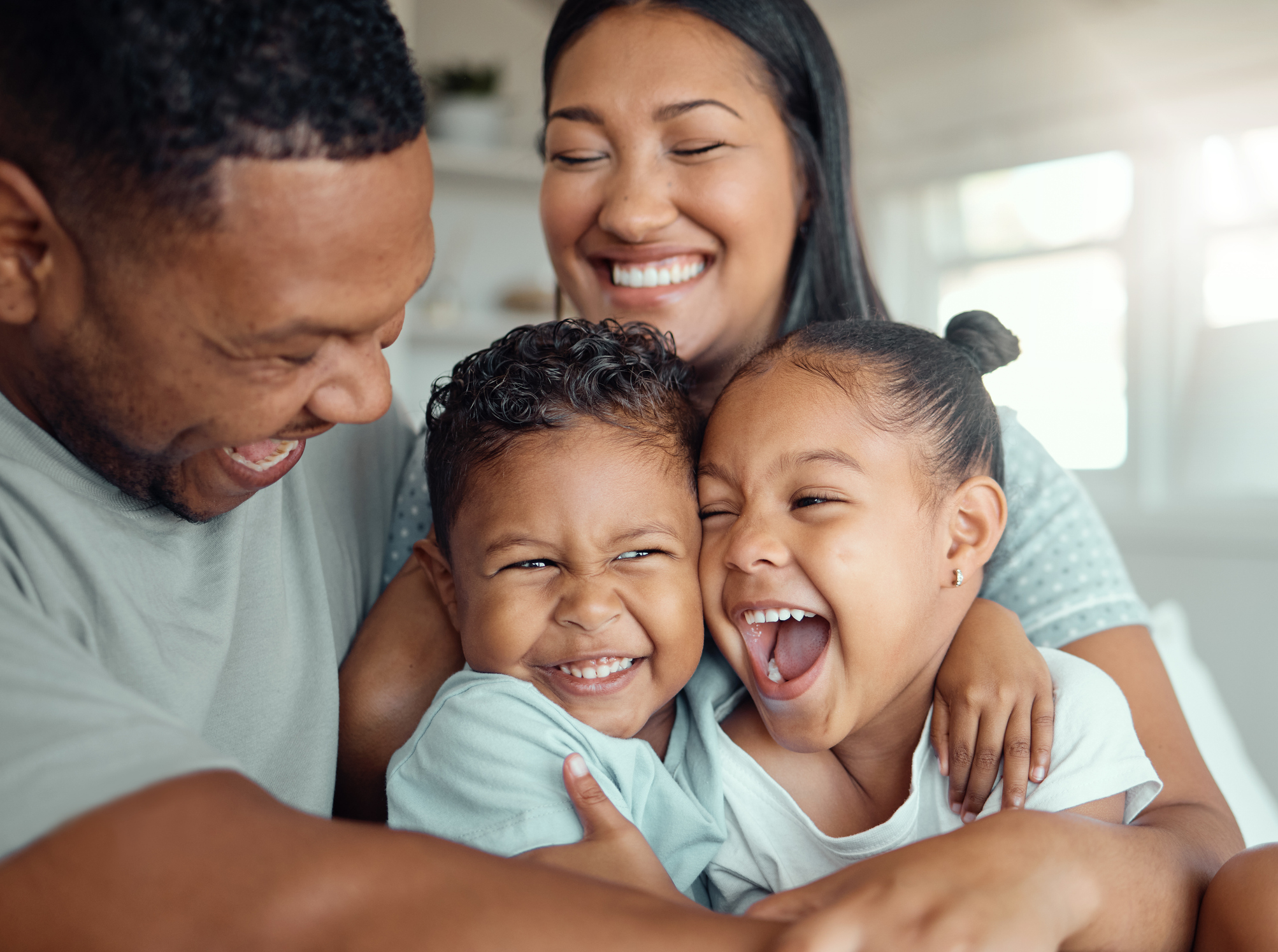 Happy funny mixed race family with two children wearing pyjamas and sitting together embracing each other at home. Cheerful parents sitting with their daughter and son laughing and having fun in the morning