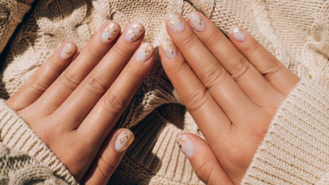 Glowing hands in sweater with knitted sleeves and with nude color manicure with gold particles on knitted background. Trendy colors of the year