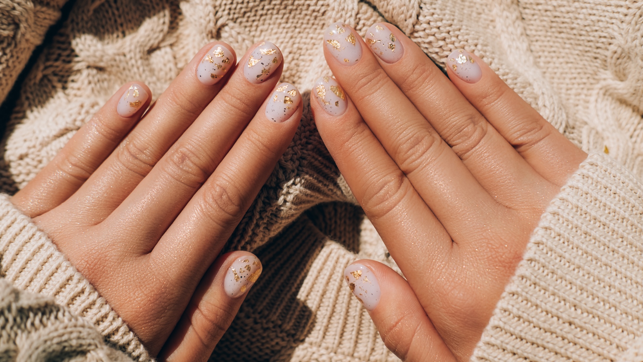 Glowing hands in sweater with knitted sleeves and with nude color manicure with gold particles on knitted background. Trendy colors of the year