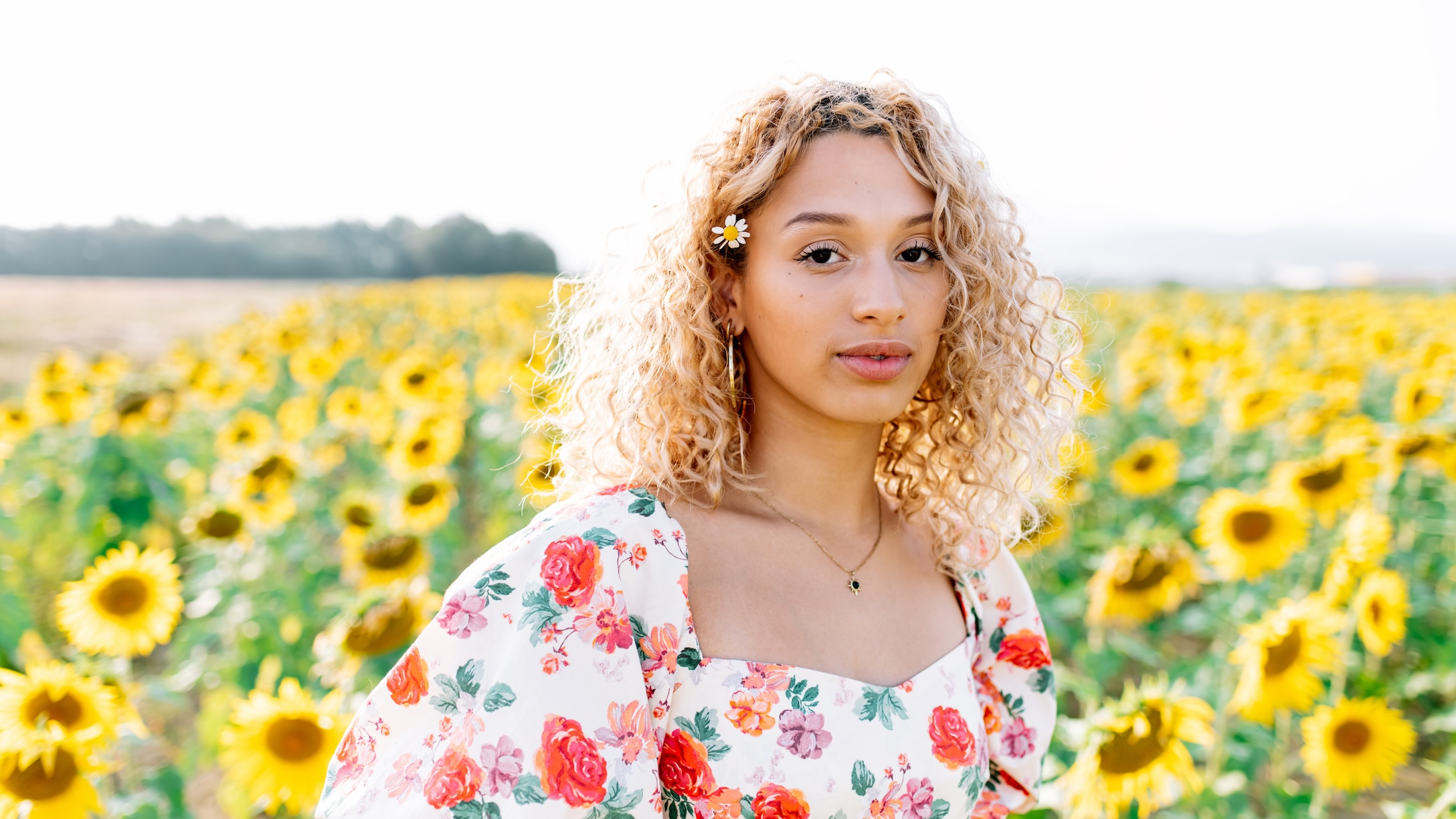 A young woman in a floral dress looking at the camera in a field of sunflowers.