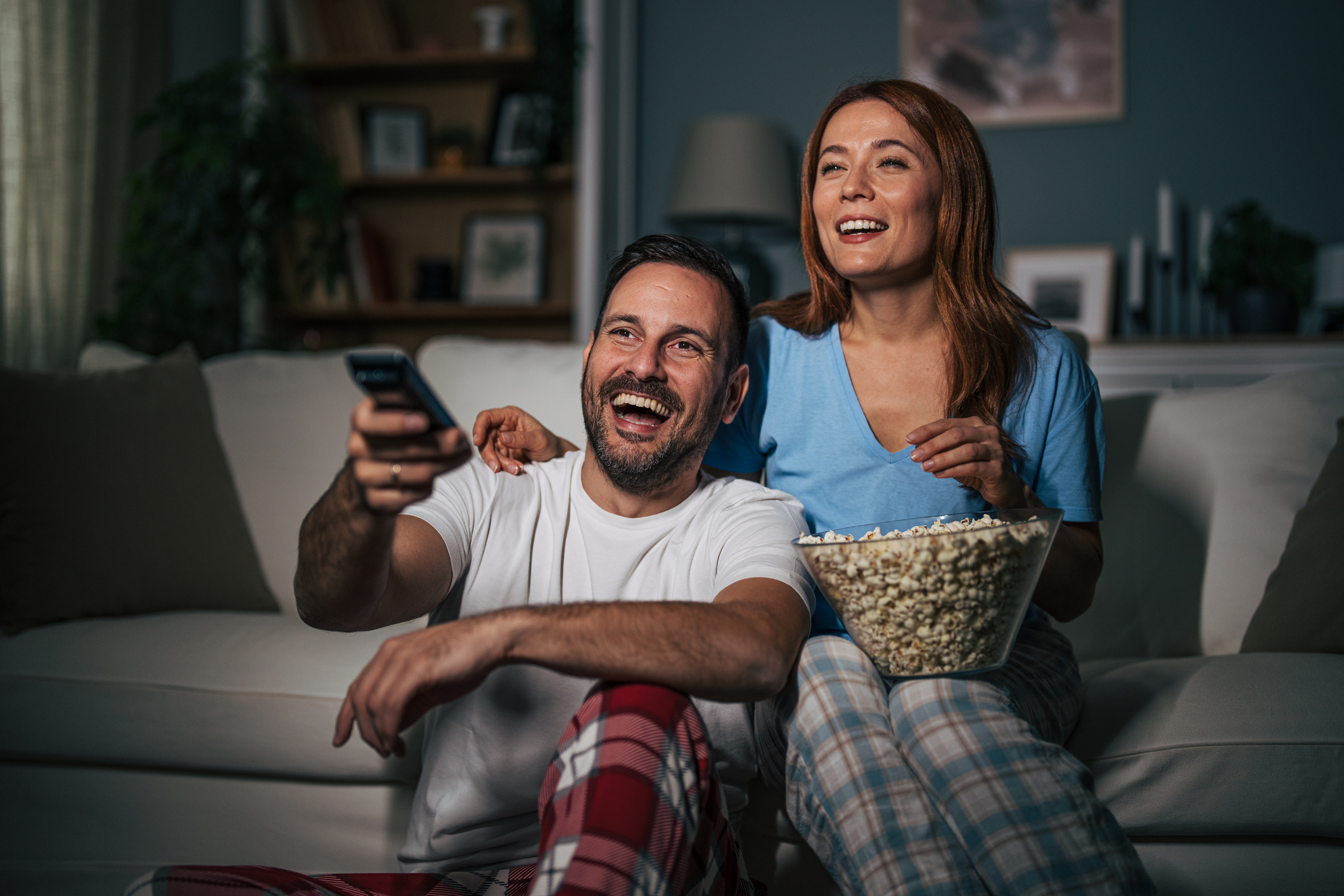 A husband and wife are watching a movie at night and eating popcorn in the living room.