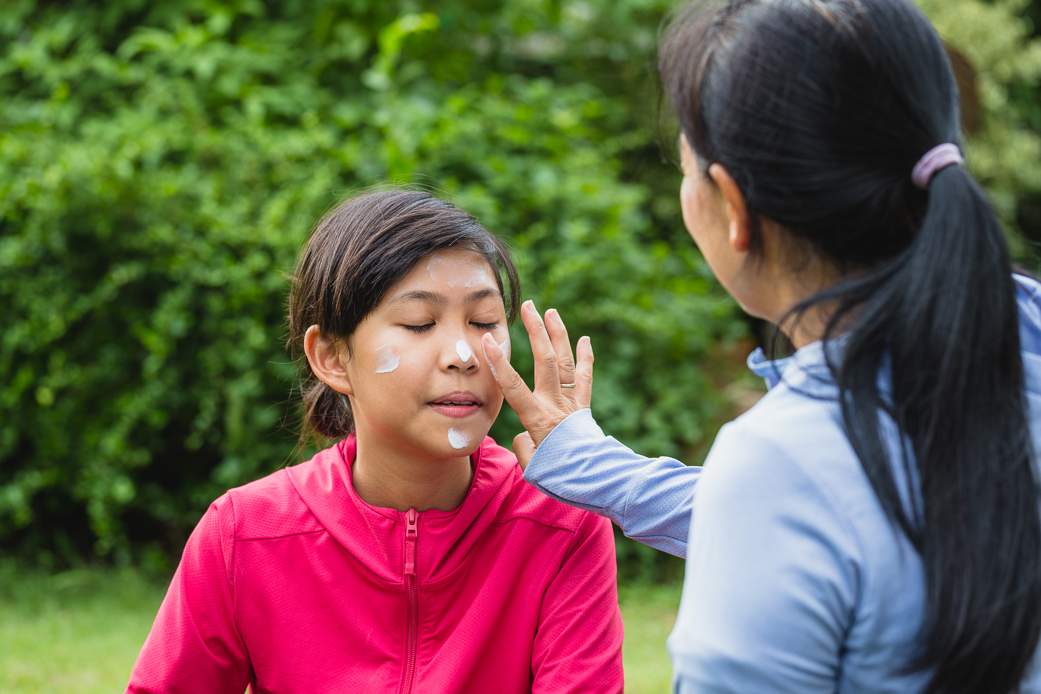 mother applying sunscreen lotion to protect her daughter's face before exercise while setting on floor in the park at outdoors