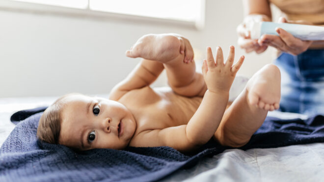 Mother changing baby's diaper in bed. Mother applies diaper rash cream.