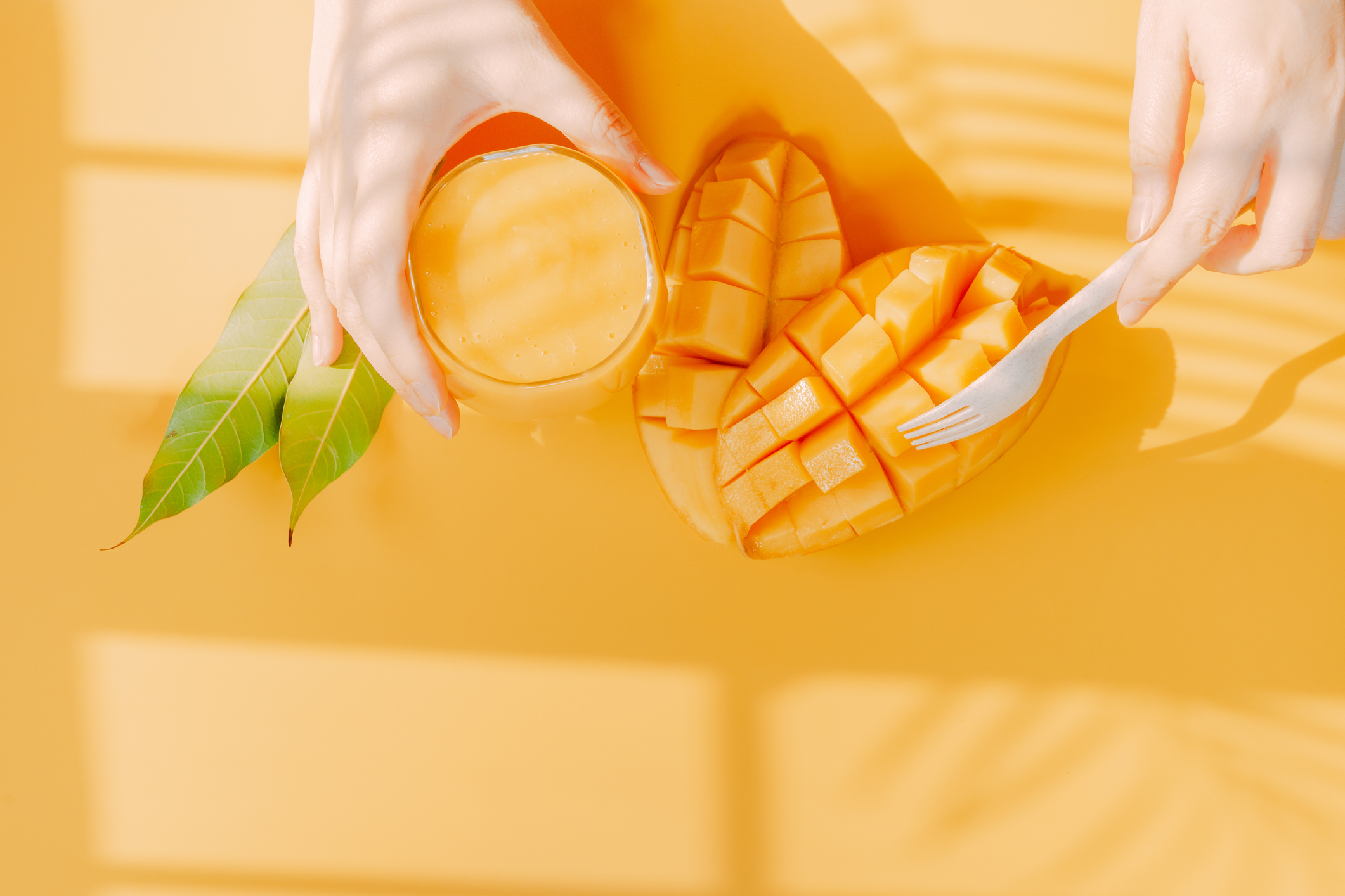 top view and flat lay summer fruit from beauty woman hand hold mango juice and use fork for eat mango slice with shadow of tropical leaf and yellow background