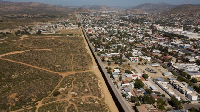 An aerial image shows steel bollard-style border wall barriers standing along the U.S.-Mexico